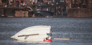 A capsized boat near an urban waterfront with a person wearing a life jacket visible in calm waters.