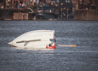 A capsized boat near an urban waterfront with a person wearing a life jacket visible in calm waters.