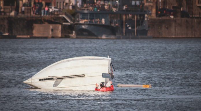 Boat Capsize Near Liberty Island Kills 2, Jersey City Crew Rescues 11 A capsized boat near an urban waterfront with a person wearing a life jacket visible in calm waters.