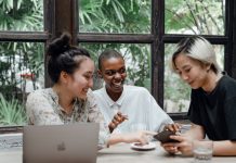 Group of cheerful friends laughing at funny meme while surfing social media on smartphone sitting at table with laptop in modern cafe