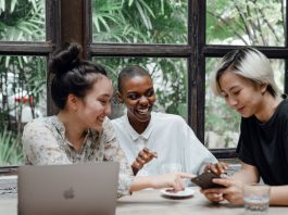 Group of cheerful friends laughing at funny meme while surfing social media on smartphone sitting at table with laptop in modern cafe