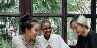 Group of cheerful friends laughing at funny meme while surfing social media on smartphone sitting at table with laptop in modern cafe