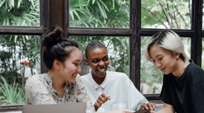 Group of cheerful friends laughing at funny meme while surfing social media on smartphone sitting at table with laptop in modern cafe