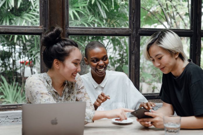 Group of cheerful friends laughing at funny meme while surfing social media on smartphone sitting at table with laptop in modern cafe