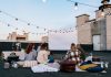 Friends enjoying a cozy evening movie on a rooftop with string lights and blankets.