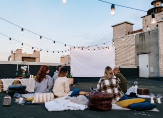 Friends enjoying a cozy evening movie on a rooftop with string lights and blankets.