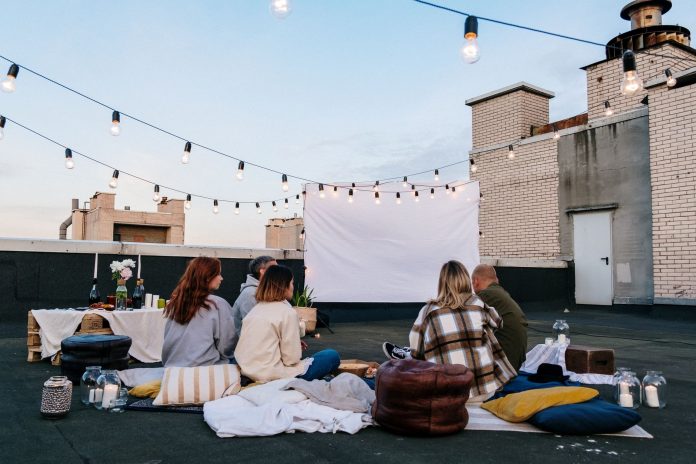 Friends enjoying a cozy evening movie on a rooftop with string lights and blankets.
