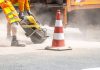 A construction worker cutting asphalt with a large saw next to a traffic cone on a street.