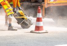 A construction worker cutting asphalt with a large saw next to a traffic cone on a street.