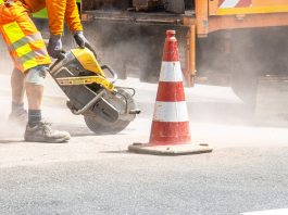A construction worker cutting asphalt with a large saw next to a traffic cone on a street.