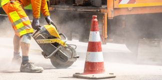A construction worker cutting asphalt with a large saw next to a traffic cone on a street.