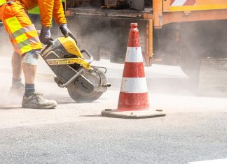A construction worker cutting asphalt with a large saw next to a traffic cone on a street.