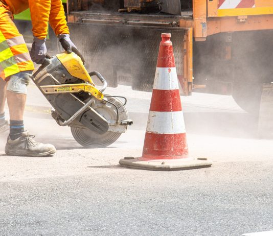 A construction worker cutting asphalt with a large saw next to a traffic cone on a street.
