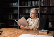 Elegant senior woman reading a manuscript at a wooden desk in a cozy library setting, surrounded by bookshelves.