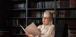 Elegant senior woman reading a manuscript at a wooden desk in a cozy library setting, surrounded by bookshelves.