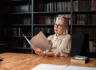 Elegant senior woman reading a manuscript at a wooden desk in a cozy library setting, surrounded by bookshelves.