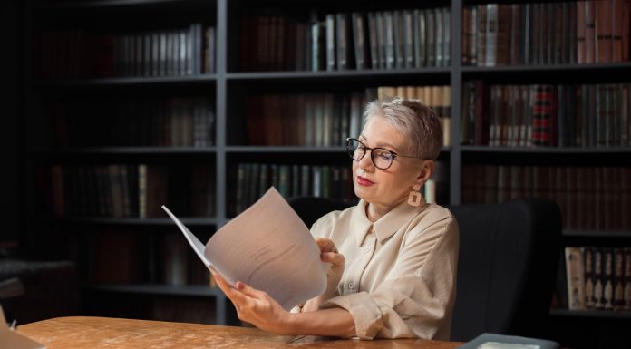 Elegant senior woman reading a manuscript at a wooden desk in a cozy library setting, surrounded by bookshelves.