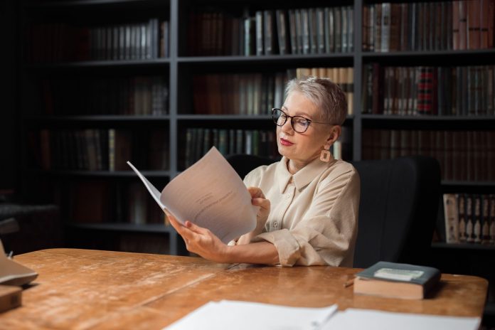 breaking-jersey-city-library-director-announces-re Elegant senior woman reading a manuscript at a wooden desk in a cozy library setting, surrounded by bookshelves.