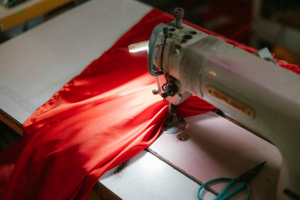 Close-up of an industrial sewing machine stitching bright red fabric, highlighted under a spotlight.