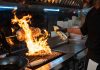 Chef uses a culinary torch to ignite a flaming grill indoors, showcasing dynamic cooking.