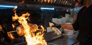 Chef uses a culinary torch to ignite a flaming grill indoors, showcasing dynamic cooking.