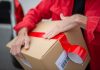 A courier in red uniform sealing a cardboard package with red tape for delivery.