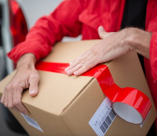 A courier in red uniform sealing a cardboard package with red tape for delivery.
