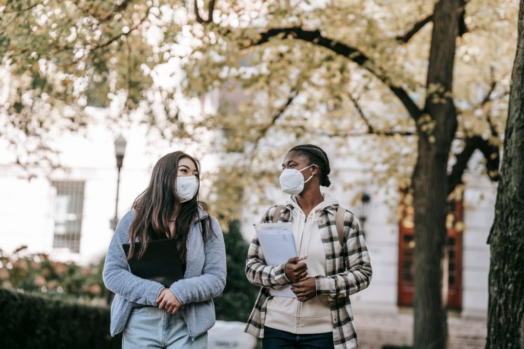 Low angle of diverse classmates in masks with folders talking about university issues while walking in park together