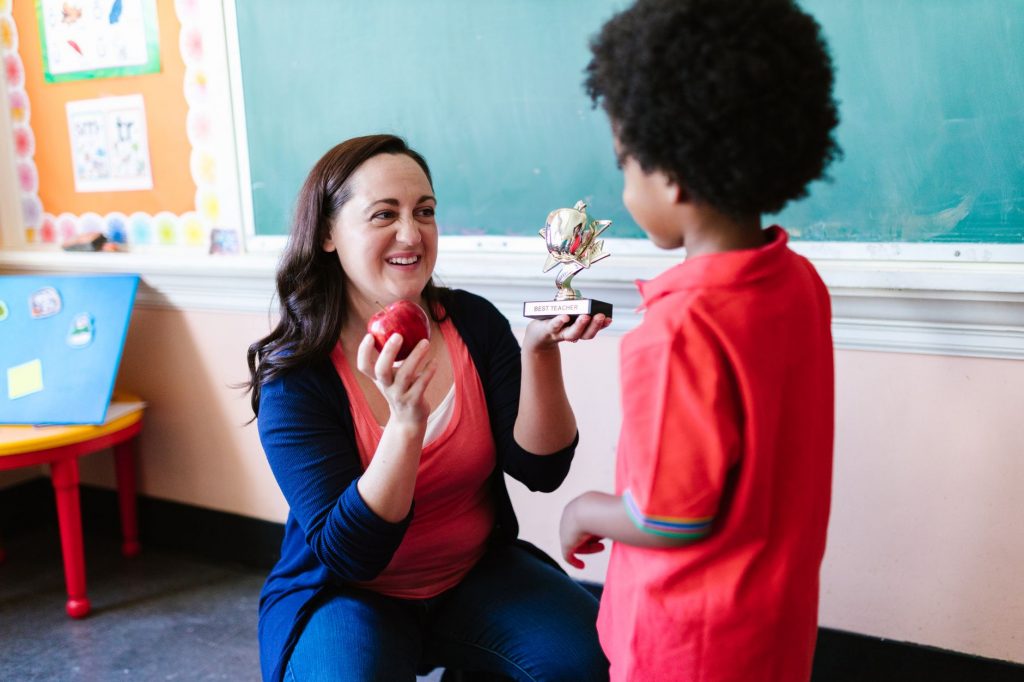 A teacher holding an apple and a student holding a trophy in a classroom setting.