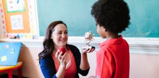 A teacher holding an apple and a student holding a trophy in a classroom setting.