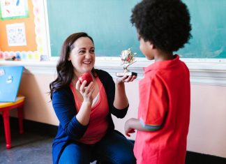 A teacher holding an apple and a student holding a trophy in a classroom setting.