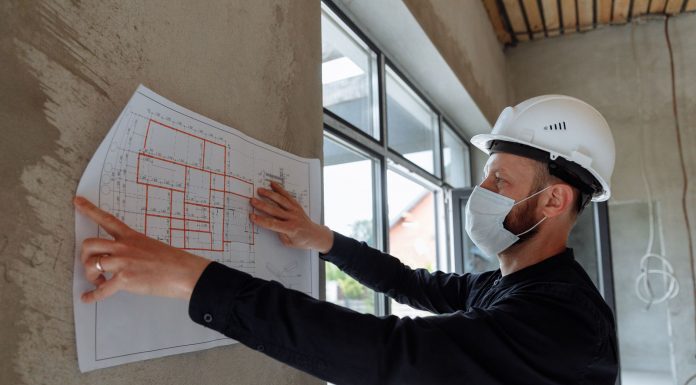 Architect wearing hard hat and mask reviews floor plans on site for new construction project.