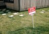 A red 'House for Rent' sign stands on a grassy lawn beside a wooden house exterior.