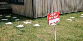 A red 'House for Rent' sign stands on a grassy lawn beside a wooden house exterior.