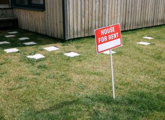 A red 'House for Rent' sign stands on a grassy lawn beside a wooden house exterior.