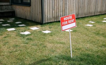 A red 'House for Rent' sign stands on a grassy lawn beside a wooden house exterior.