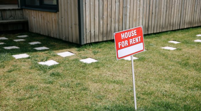 A red 'House for Rent' sign stands on a grassy lawn beside a wooden house exterior.