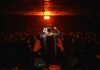 Back view of a performer engaging a lively crowd under dramatic lighting in a dark, indoor setting.