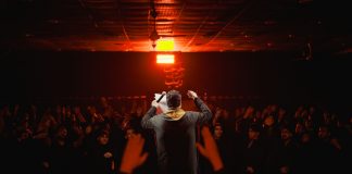 Back view of a performer engaging a lively crowd under dramatic lighting in a dark, indoor setting.