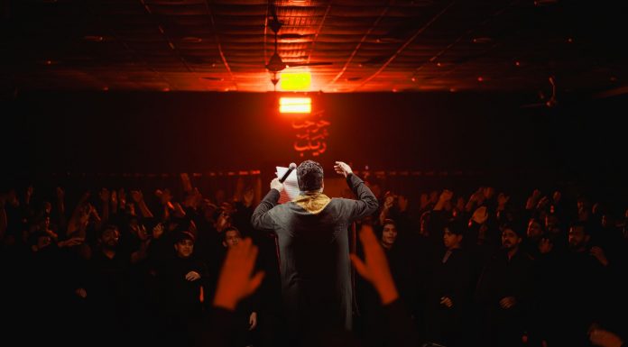 Back view of a performer engaging a lively crowd under dramatic lighting in a dark, indoor setting.