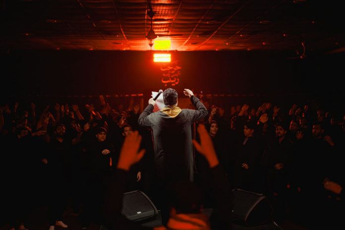 Back view of a performer engaging a lively crowd under dramatic lighting in a dark, indoor setting.