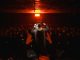 Back view of a performer engaging a lively crowd under dramatic lighting in a dark, indoor setting.