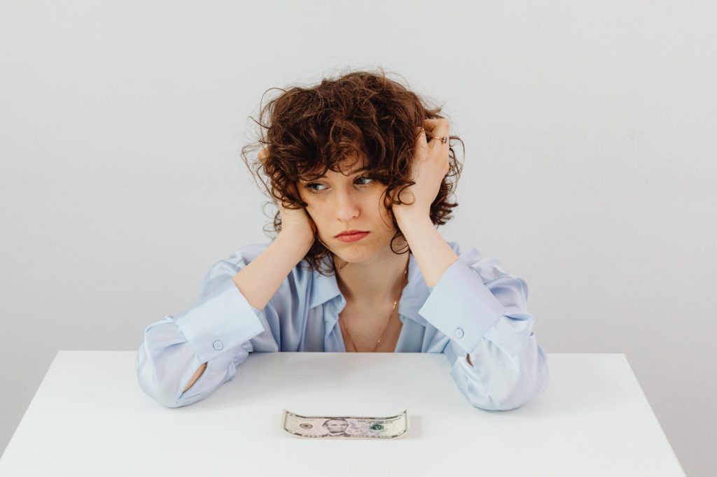 A woman in a blue blouse looks worried with a dollar bill on a table, symbolizing financial stress.