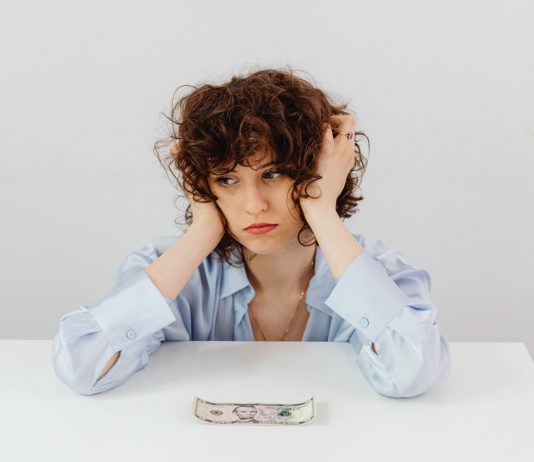A woman in a blue blouse looks worried with a dollar bill on a table, symbolizing financial stress.