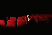Jersey City’s Cultural Affairs Director Steps Down After 25 Years A woman sitting alone surrounded by empty red theater seats in a dark auditorium.