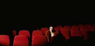 A woman sitting alone surrounded by empty red theater seats in a dark auditorium.