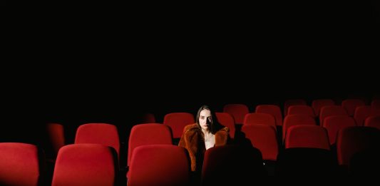 A woman sitting alone surrounded by empty red theater seats in a dark auditorium.