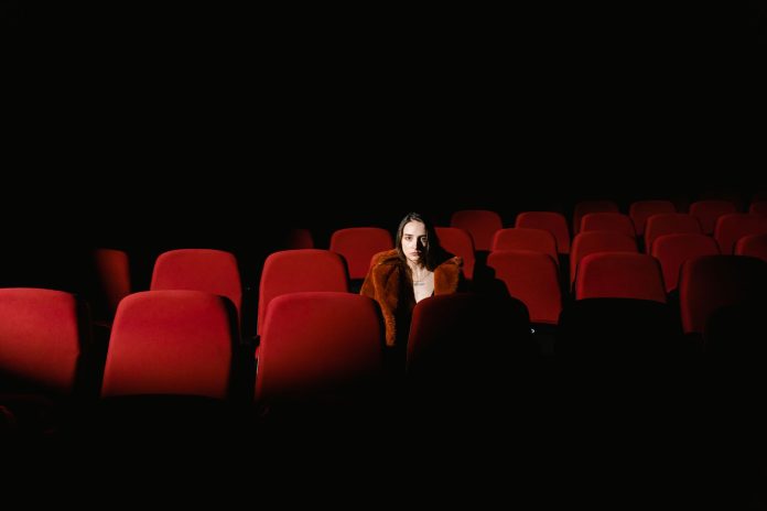 A woman sitting alone surrounded by empty red theater seats in a dark auditorium.