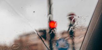 Blurred red traffic light seen through raindrops on a car windshield in a city setting.