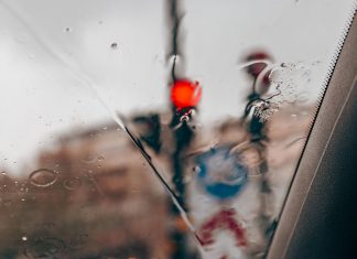 Blurred red traffic light seen through raindrops on a car windshield in a city setting.